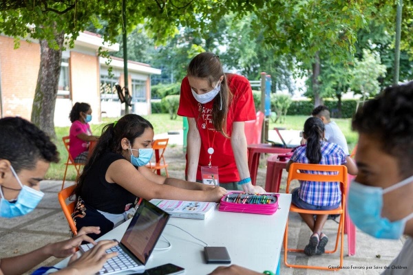 operatrice svae the children con dei bambini, seduti a un tavolo esterno mentre fanno attività educative, tutti con mascherina per proteggersi dal covid