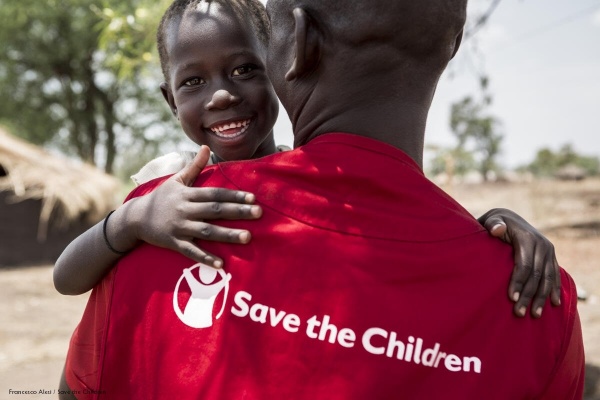 operatore save the children di spalle con maglietta rossa e logo con in braccio un bambino sorridente