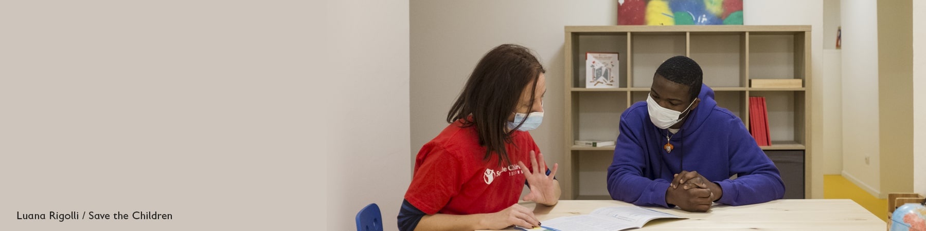seduti a un tavolo, indossando le mascherine, un operatrice di Save the Children con maglietta rossa e un ragazzo con maglioncino blu guardano insieme degli esercizi su un foglio