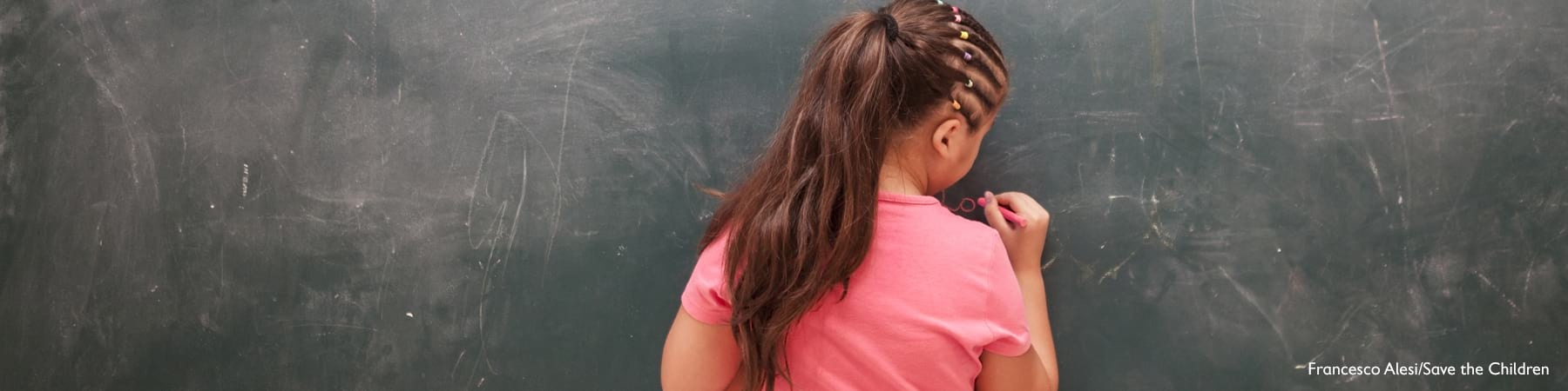 Bambina di spalle con maglietta rosa scrive su una lavagna