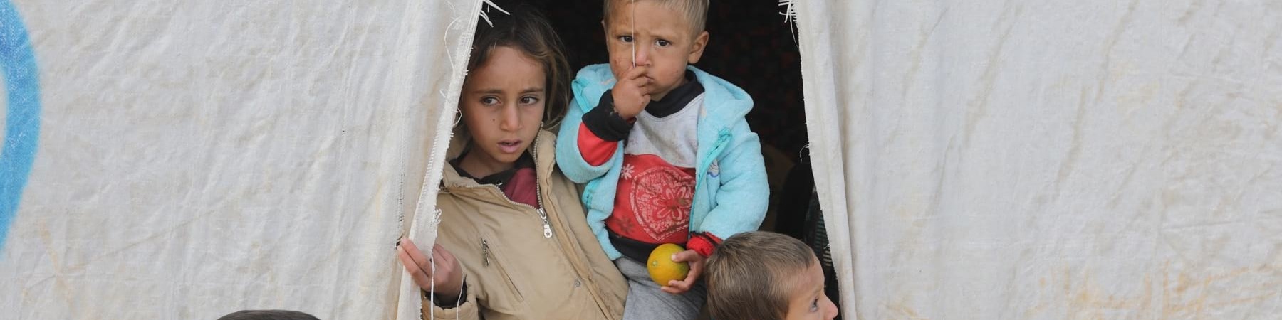 quattro bambini siriani sono sulla soglia di una tenda di un campo rifugiati, una di loro è più grande e tiene in braccio un altro.