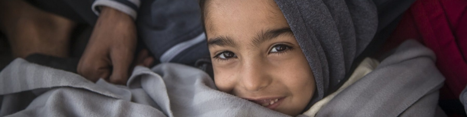 Un bambino con cappuccio grigio in testa sorride avvolto in una coperta anche essa grigia.