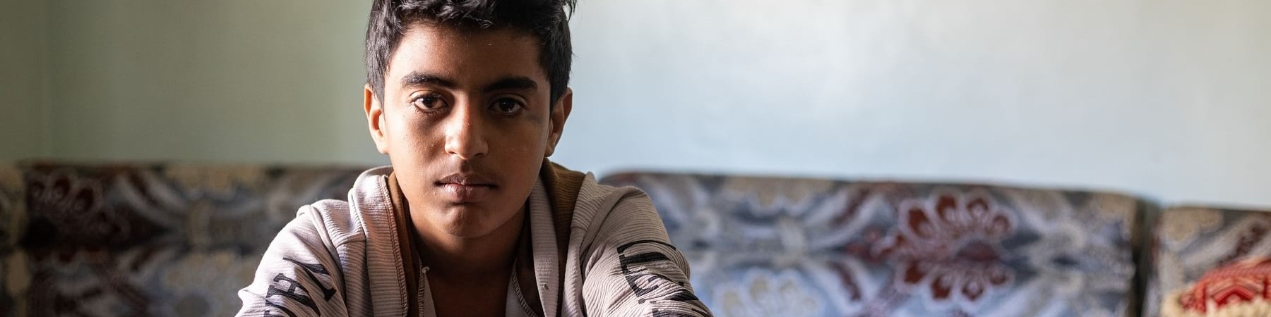 bambino yemenita seduto tiene le braccia appoggiate sulle gambe incrociate. Indossa maglietta beige e jeans