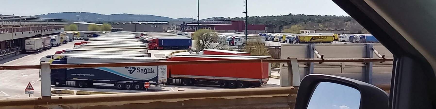 piazzale di camion ripreso dal finestrino di una macchina della quale si vede anche uno specchietto.