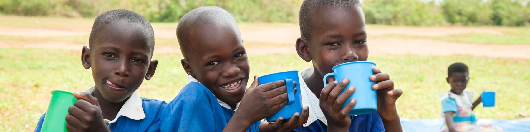 tre bambini ugandesi vestiti di blu, seduti su un ramo bevono acqua da alcune tazze colorate