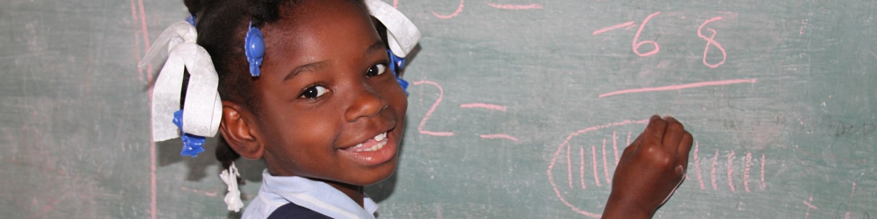 bimba che scrive alla lavagna a scuola e ci guarda con un grande sorriso. Vestita con una camicia azzurra ha un nastro tra i capelli pettinati con due treccine.