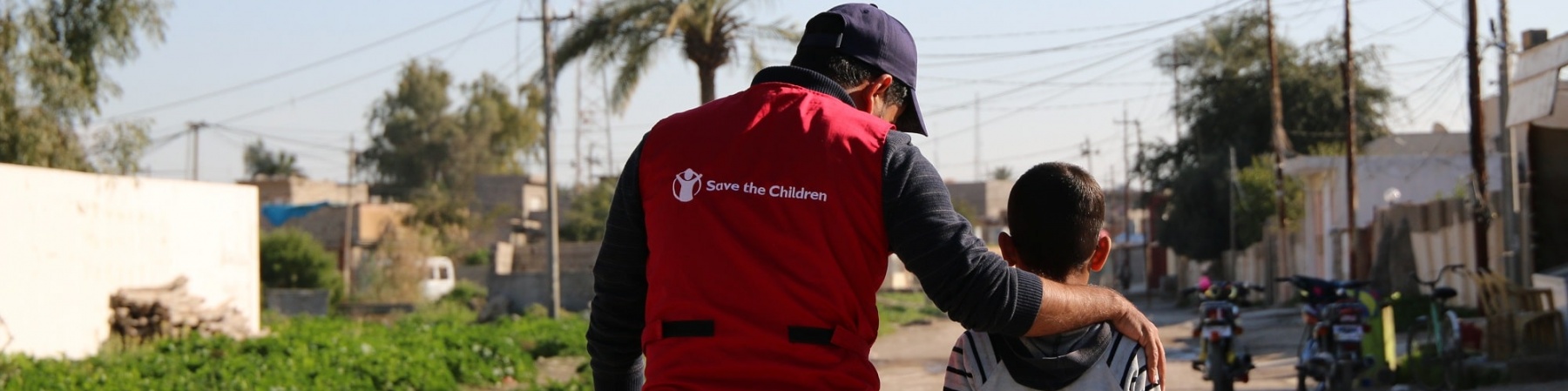 operatore save the children con maglietta rossa cammina al fianco di un bambino, entrambi li vediamo di spalle 