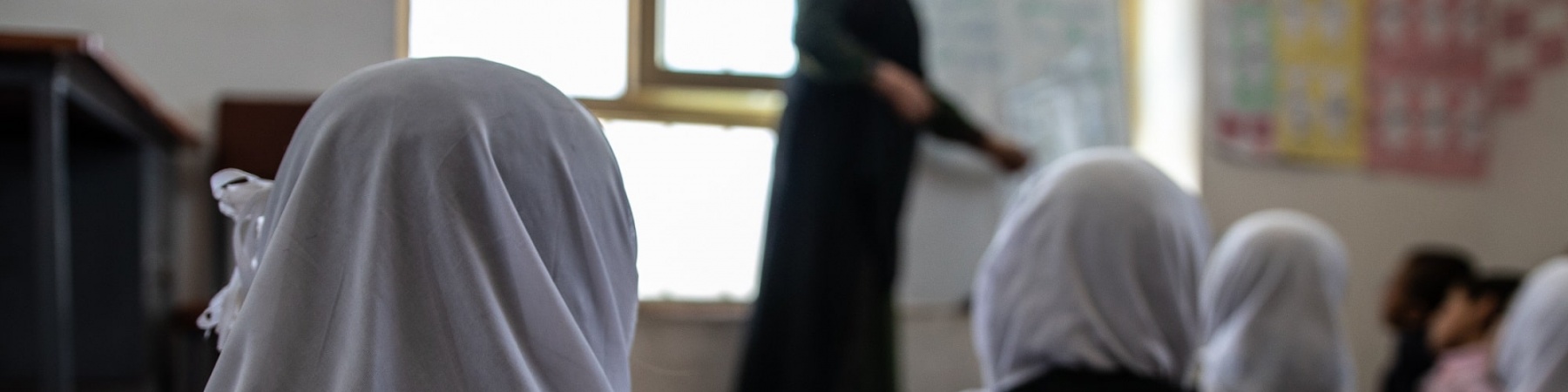 ragazze di spalle, sedute per terra in un aula scolastica, indossano un velo bianco e guardano verso la maestra