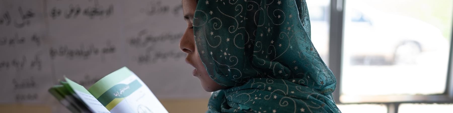 Una bambina afghana di profilo con vestito e velo verde ricamato tiene in mano davanti a sé un libro aperto e lo legge