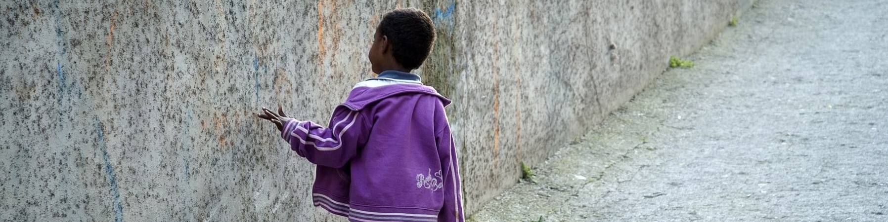 Bambino migrante cammina di spalle toccando muro grigio