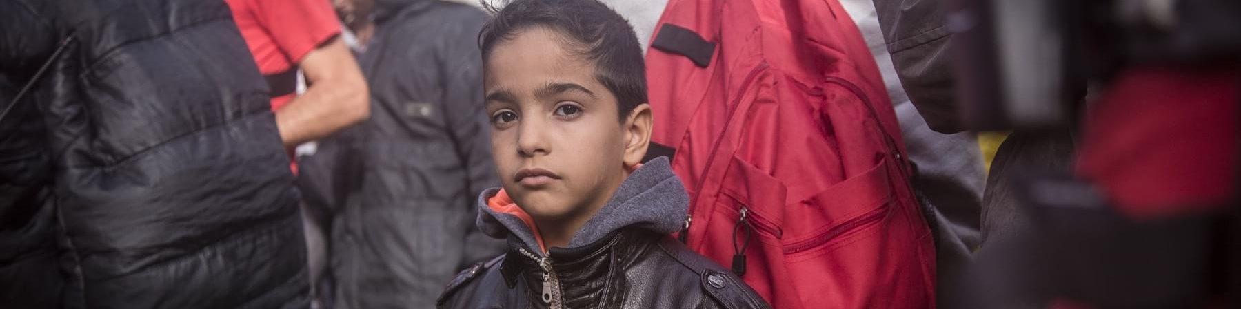 Un bambino in mezzo a tante persone guarda in camera. Indossa una giacca di pelle nera con la zip. Lui è moro e ha i capelli corti