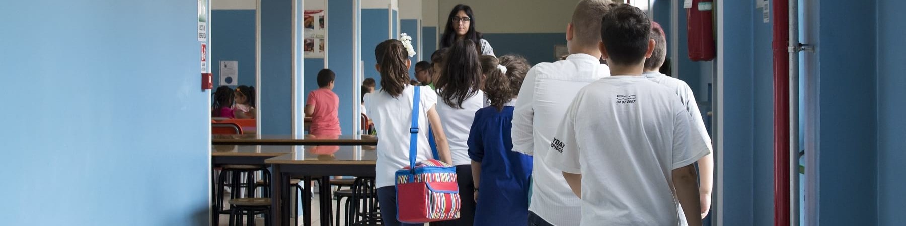 Bambini in fila nel corridoio di una scuola sono inquadrati di spalle mentre camminano verso l insegnate sullo sfondo girata verso di loro