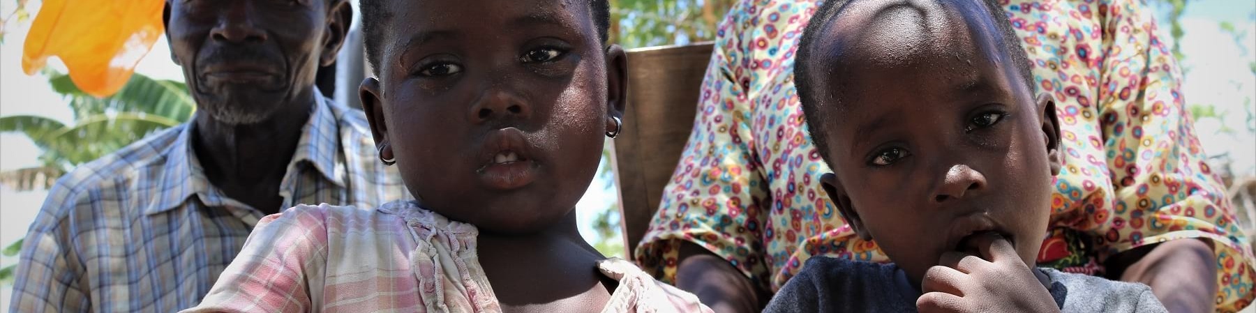 Due bambini del Mozambico in primo piano e dietro di loro un uomo e una donna guardano in camera ripresi leggermente dal basso. Uno dei bambini ha un dito in bocca e una maglietta nera, l altra ha un vestitino bianco