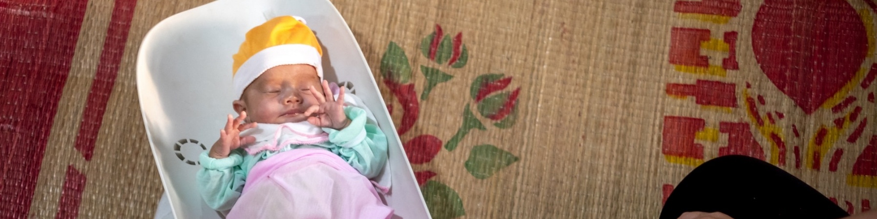 neonato in una vaschetta di plastica per pesarlo, vestito con vestitini caldi e colorati