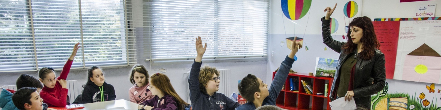 bambini in classe con maestra che alzano la mano