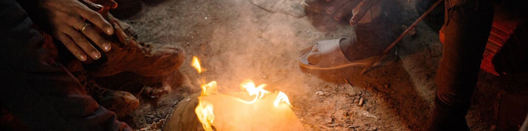 Focolare acceso a terra intorno al quale si scaldano mani di persone. Non sono visibili volti e corpi ma solo le braccia che si protendono verso il fuoco