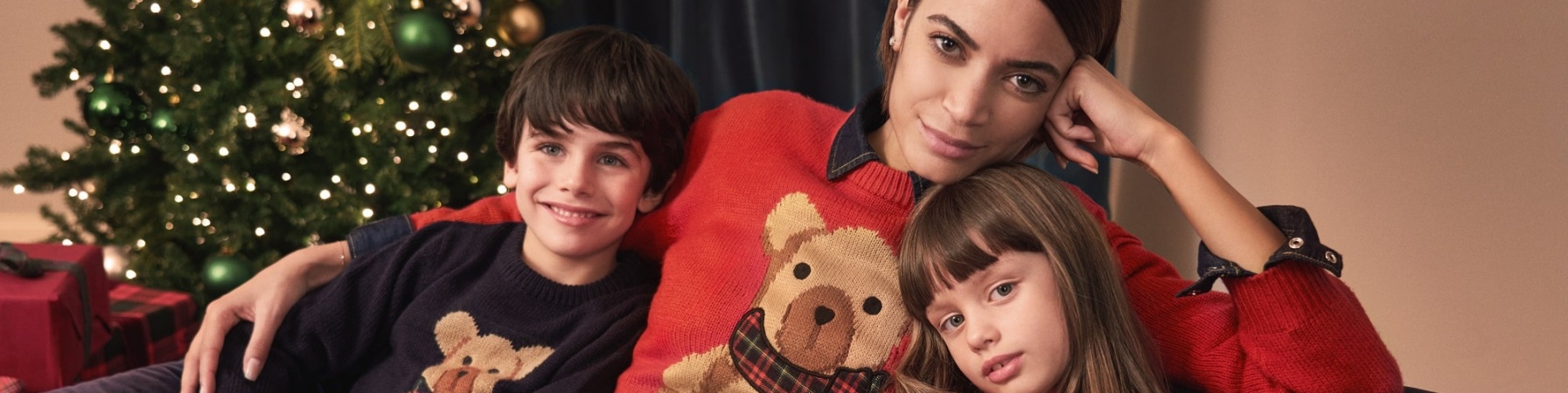 la cantante Elodie seduta su un divano insieme a una bambina e un bambino. Tutti e 3 indossano un maglione natalizio e alle loro spalle cè un albero di Natale