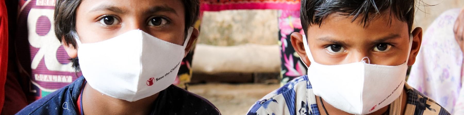 Due bambini indiani seduti vicini indossano mascherina bianca save the children. Uno ha una camicia a righe, l altro una felpa blu