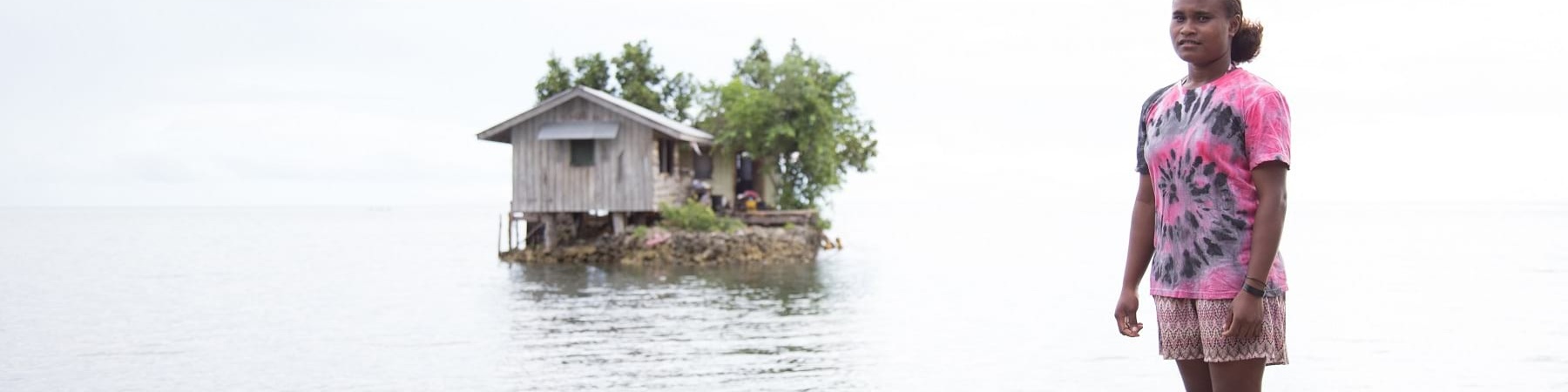 Una ragazza delle Solomon Island è in piedi sulla riva di un lago e la sua casa è un isolotto artificiale alle sue spalle