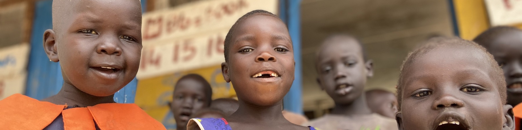 Tre bambini ugandesi in primo piano sorridono in camera. Sullo sfondo struttura in legno della scuola