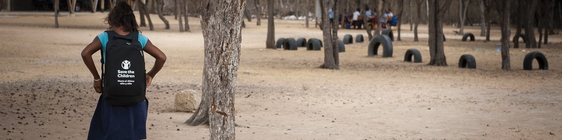bambina di spalle con lo zaino cammina su terreno vicino a un albero