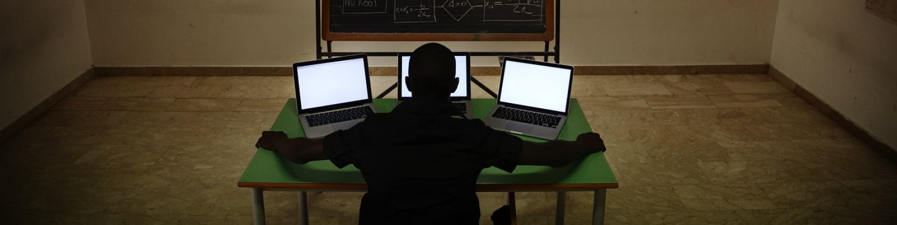 Ragazzo di spalle seduto al tavolo con tre computer