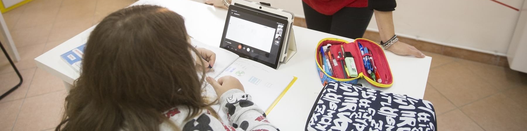 una bambina di spalle seduta a un tavolo davanti a un tablet. Di fronte e lei in piedi con le braccia poggiate al tavolo una operatrice save the children