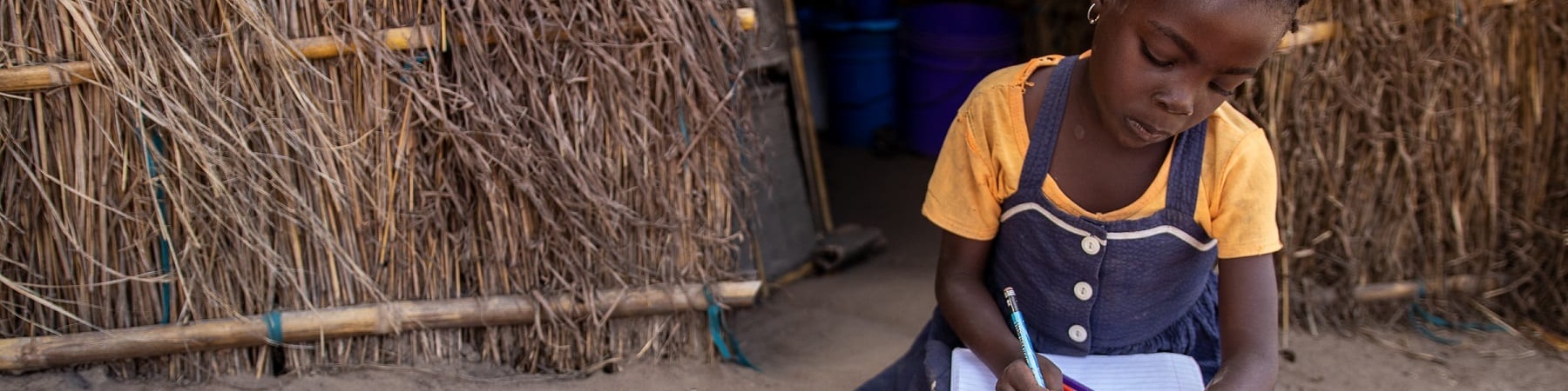 una bambina africana con maglietta gialla e vestito jeans è seduta a terra accanto alla sua capanna mentre è intenta a scrivere qualcosa sopra un quaderno