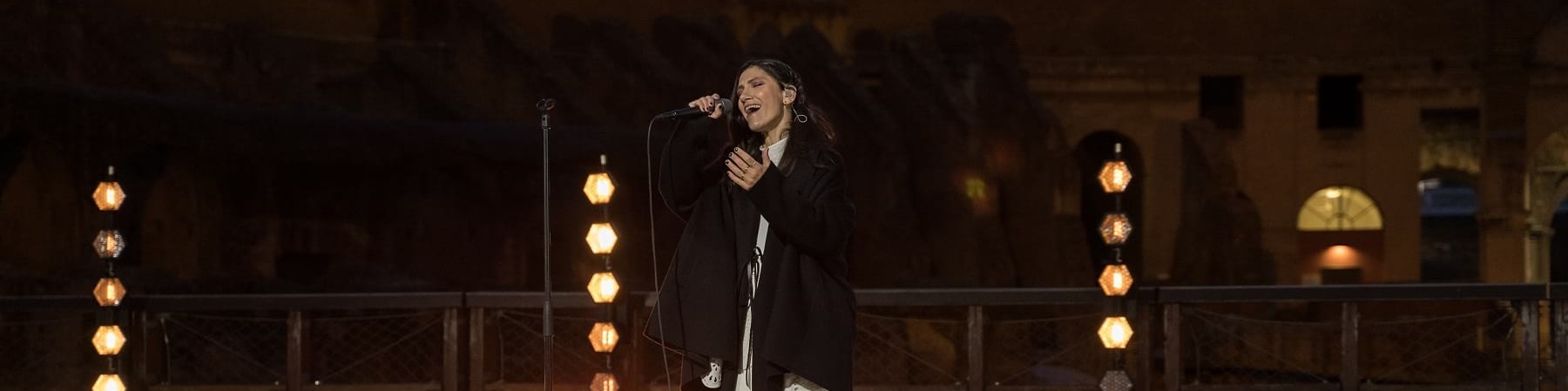 La cantautrice Elisa canta in piedi con microfono all interno del Colosseo circondata da candele accese sul palco. Elisa indossa un vestito lungo bianco e una giacca lunga nera