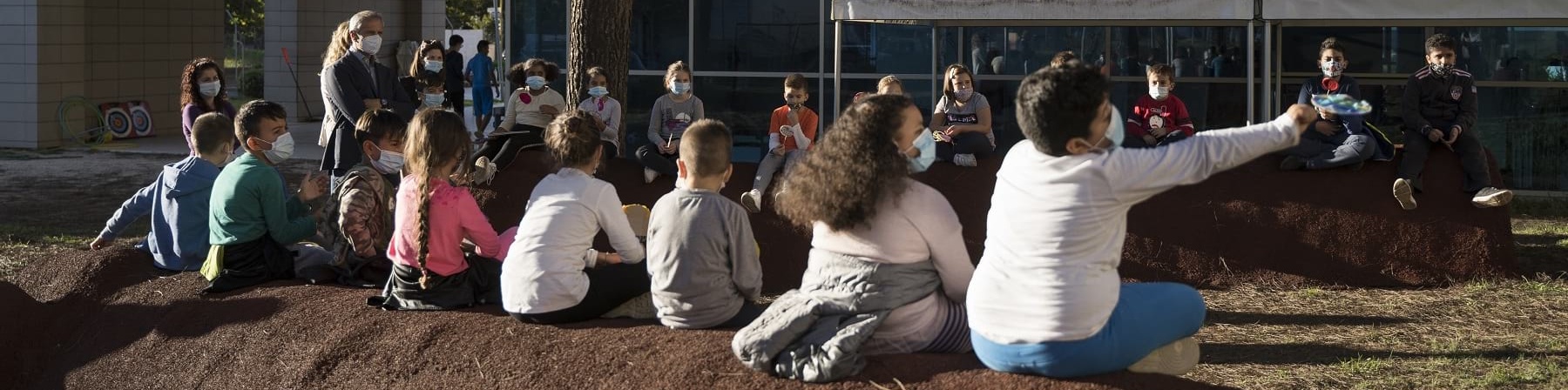 Foto all aperto, una fila di bambini di spalle seduti su un terrapieno guardano due gazebo bianchi montati di fronte a loro