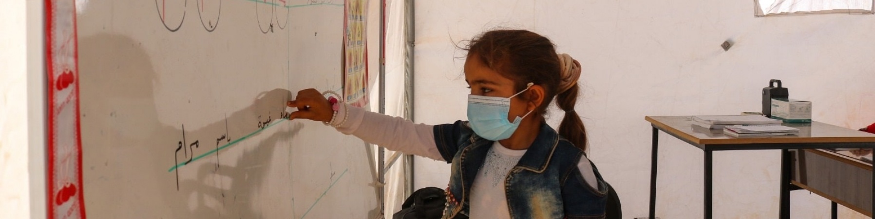 bambina scrive su una lavagna bianca in un aula, indossa una mascherina chirurgica per proteggersi dal Covid-19