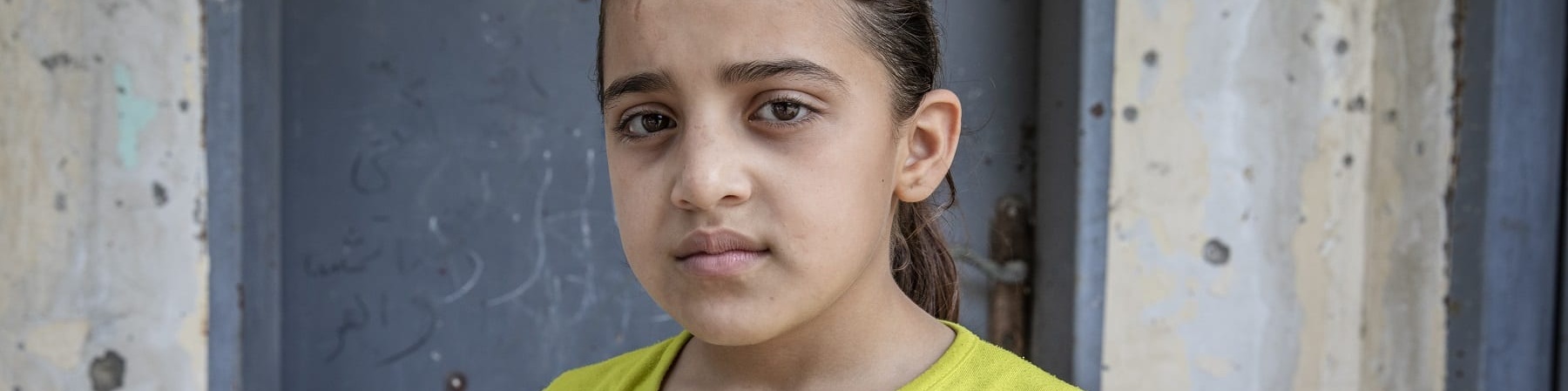 Mezzo busto di una bambina palestinese che indossa una maglietta gialla a maniche corte con alcune scritte colorate sopra. La bambina è castana ed ha i capelli raccolti in una coda.