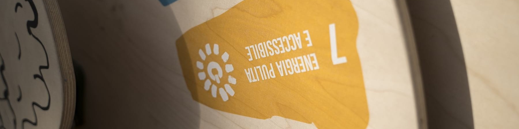 Una ruota di legno appesa a un muro mostra in blocchi colorati gli obiettivi di sviluppo sostenibile. Nella foto viene inquadrato prevalentemente il goal "energia pulita e accessibile" su sfondo giallo