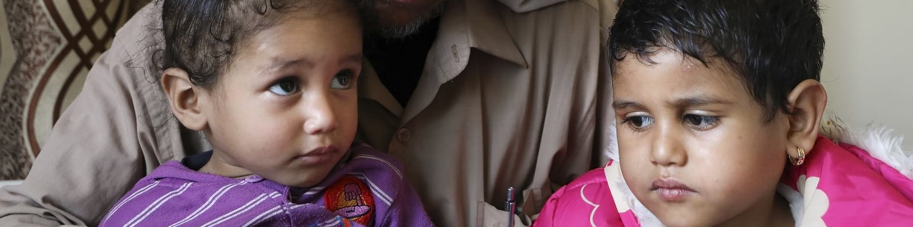 Un papà yemenita con turbante bianco in testa tiene in braccio le sue due bambine e le guarda. Entrambe hanno i capelli corti e neri, una guarda verso l alto e l altra verso la sua sorellina. Indossano magliette viola e rosa