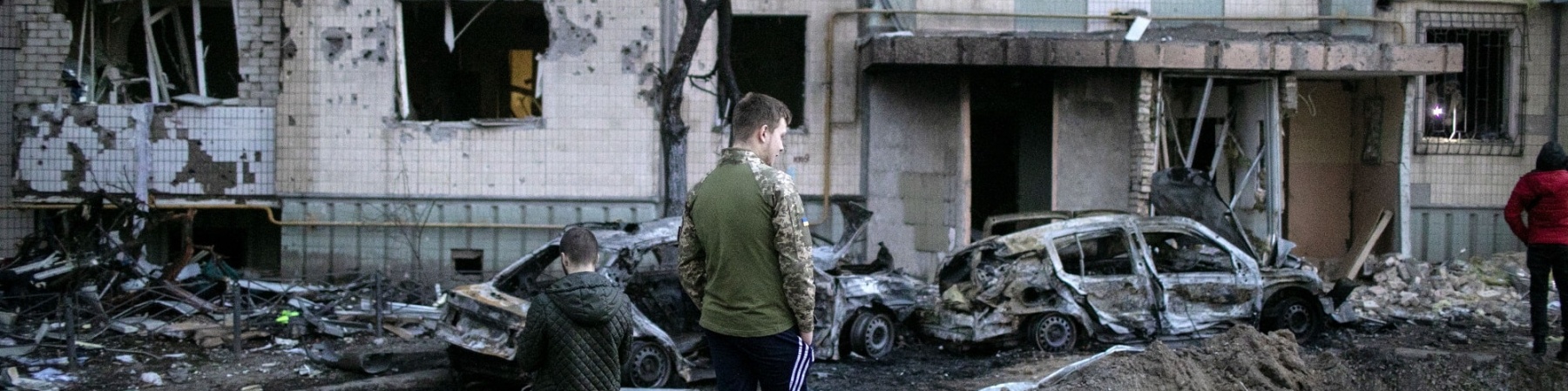 Ucraina, un bambino e un ragazzo in piedi di spalle davanti a edifici e macchine distrutti e in macerie a causa dei bombardamenti
