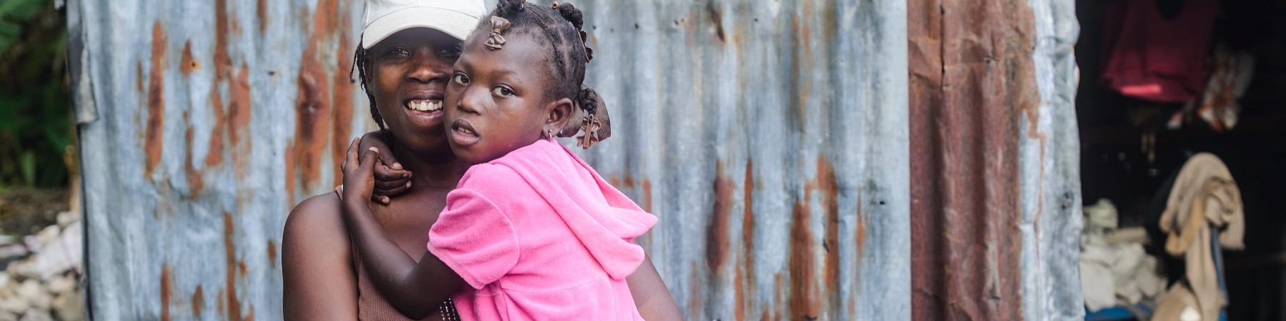 Una donna haitiana tiene in braccio la sua bambina che indossa un vestito rosa e le tiene le braccia strette intorno al collo. La donna indossa una maglietta marrone e un cappellino bianco in testa. Dietro di loro si vedono le lamiere di una baracca.