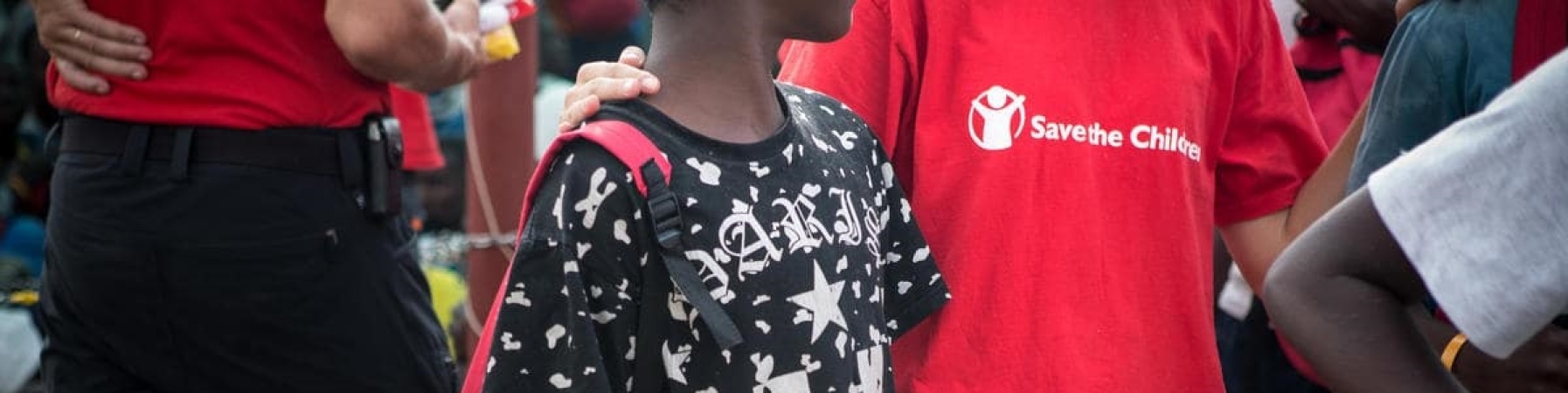 bambino con maglietta nera e zaino sulle spalle accanto ad operatore Save the Children 