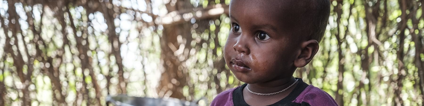 Bambino africano seduto a terra riparato in una capanna indossa una maglietta viola a strisce gialle e guarda alla sua destra.