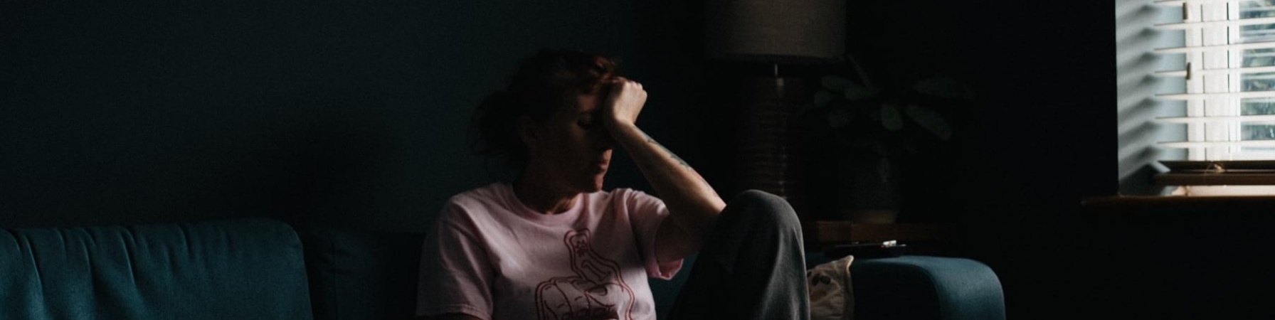 Una donna in una stanza semi buia è seduta sul divano e ha la testa appoggiata alla mano con un aria molto afflitta