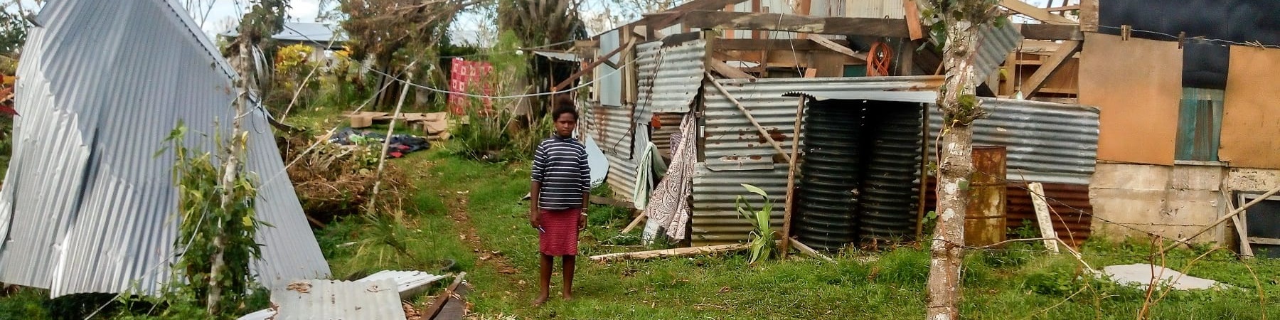 Una bambina in lontananza con gonna rossa e maglietta a righe bianche e nere si trova in piedi e scalza sul prato. Dietro di lei una casa distrutta al passaggio di un ciclone sulle isole Fiji