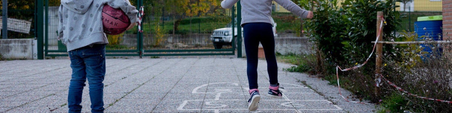 una bambina sulla sinistra tiene un pallone sotto il braccio mentre guarda l'amica che salta sui riquadri a terra del gioco campana