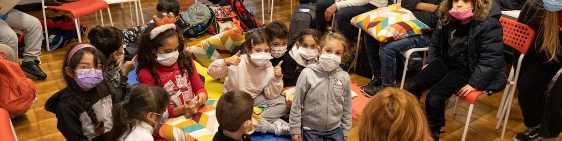 bambini e bambine seduti a terra in cerchio con mascherine che leggono un libro insieme all'operatrice