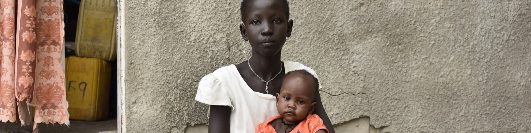 Una ragazzina sud sudanese seduta sui gradini davanti casa sua tiene in braccio una bambina piccola vestita di rosso.