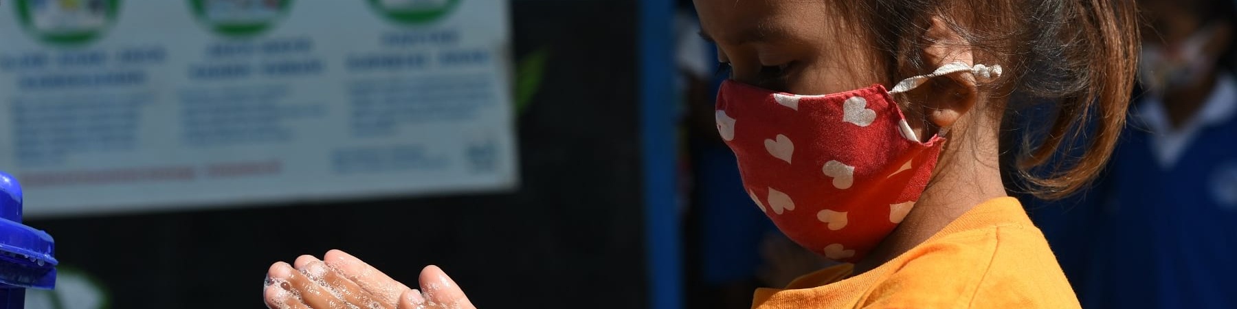 bambina indonesiana di profilo si lava le mani mentre indossa mascherina colorata sul viso.