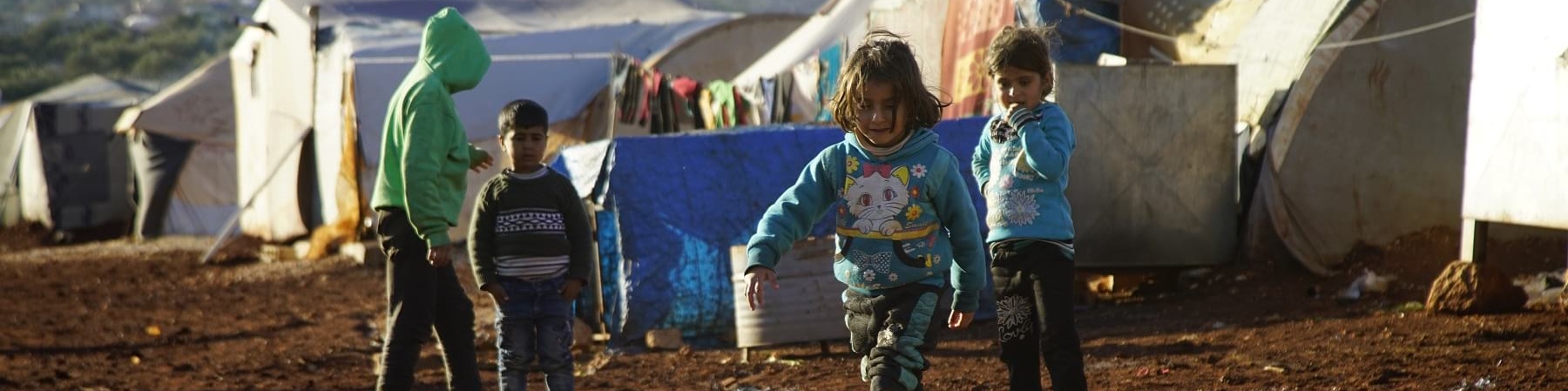 Un gruppo di bambini siriani, quattro in tutto, sono ripresi in piedi da lontano all interno di un campo profughi e su un terreno infangato a causa delle piogge. In particolare, una bambina con stivaletti rosa avanza camminando verso una grande pozzanghera d acqua di fronte a lei.