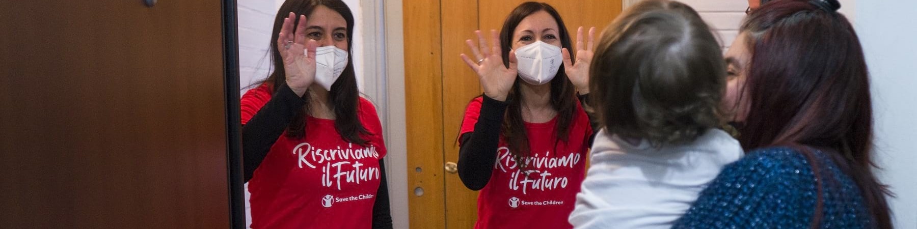 Due operatrici di Save the Children con maglietta della campagna Riscriviamo il Futuro salutano mamma e figlia davanti a loro sull uscio della porta di casa