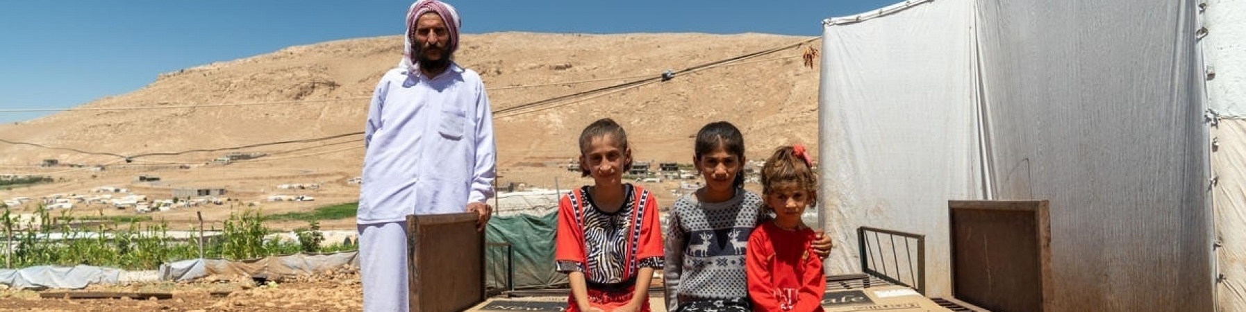 tre bambini yazidi seduti su una panca e in piedi alla loro destra il padre vestito di bianco