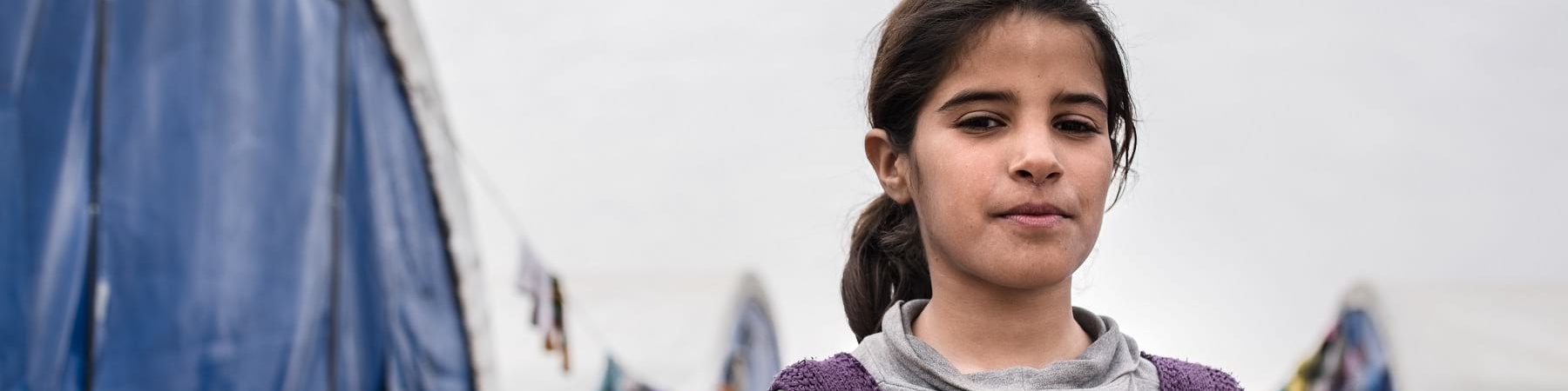bambina irachena di circa 12 anni è accanto a una tenda di un campo rifugiati, è ripresa a mezzo busto e indossa una felpa grigia e viola. I suoi capelli sono castani e legati in una coda