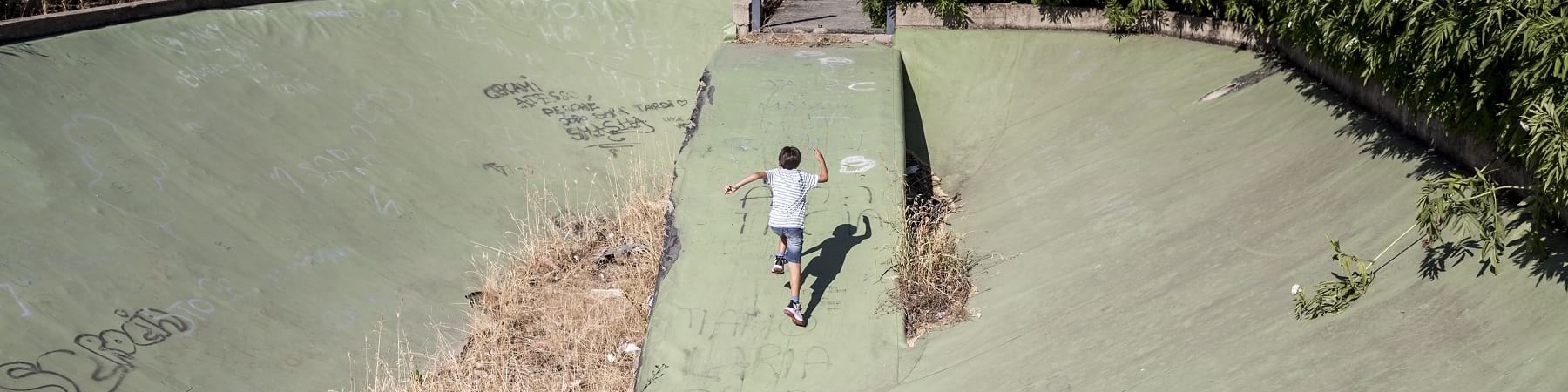 Bambino di spalle e in lontananza corre sulla salta di una pista da skate abbandonata