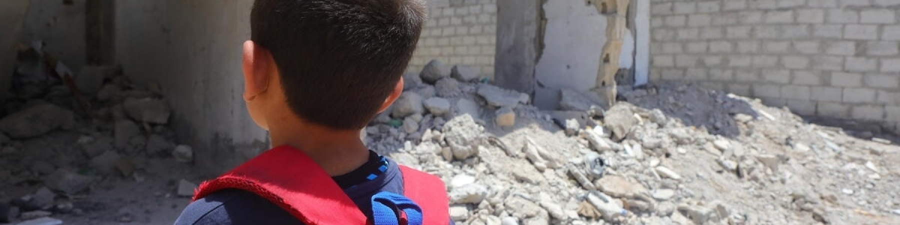 bambino con zaino rosso che guarda le macerie di una scuola distrutta 